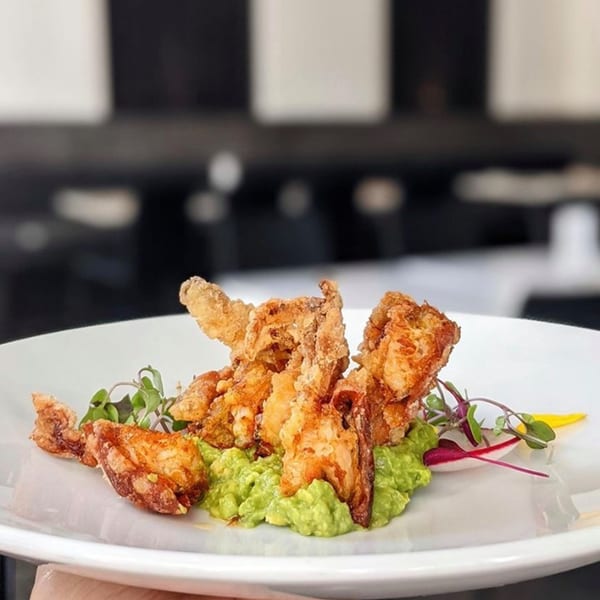 Crispy fried soft-shell crab tempura with guacamole, microgreens, and radish on a white plate.